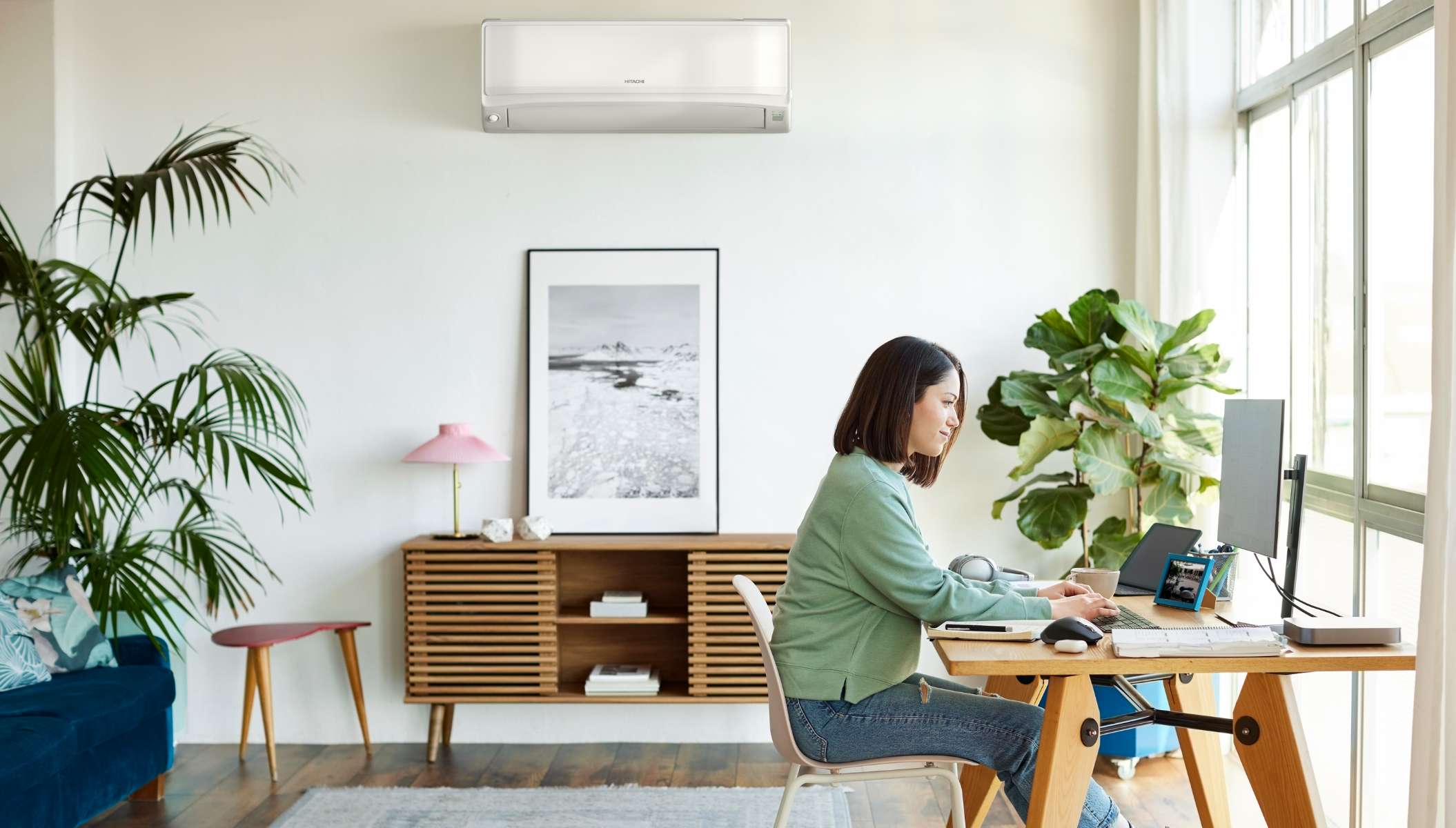 A modern, bright living room with a woman working remotely in front of a window. An indoor air conditioning unit is installed on the wall, ensuring a comfortable environment.