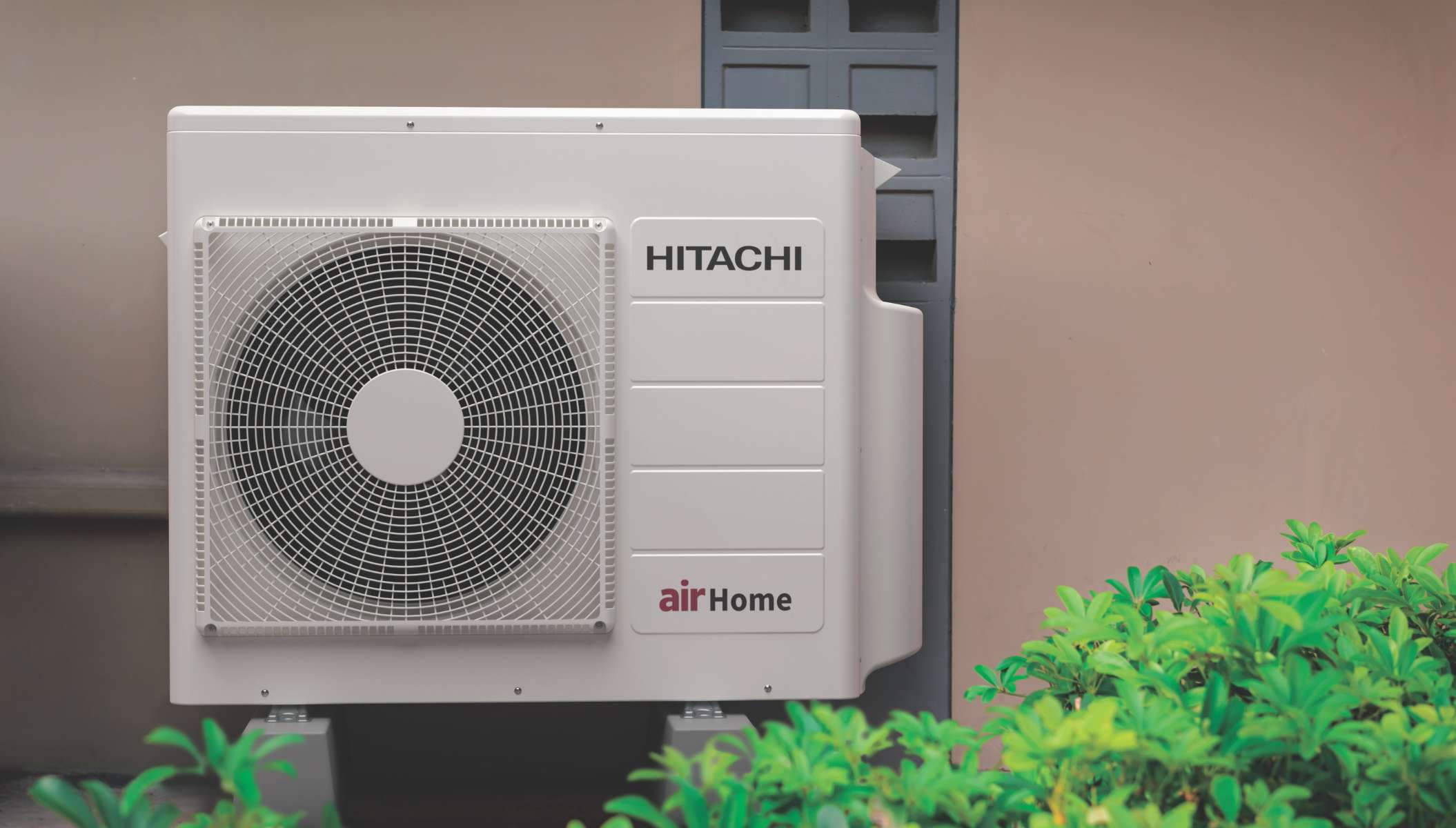 An outdoor air conditioning unit installed against a beige wall with green plants in the foreground.