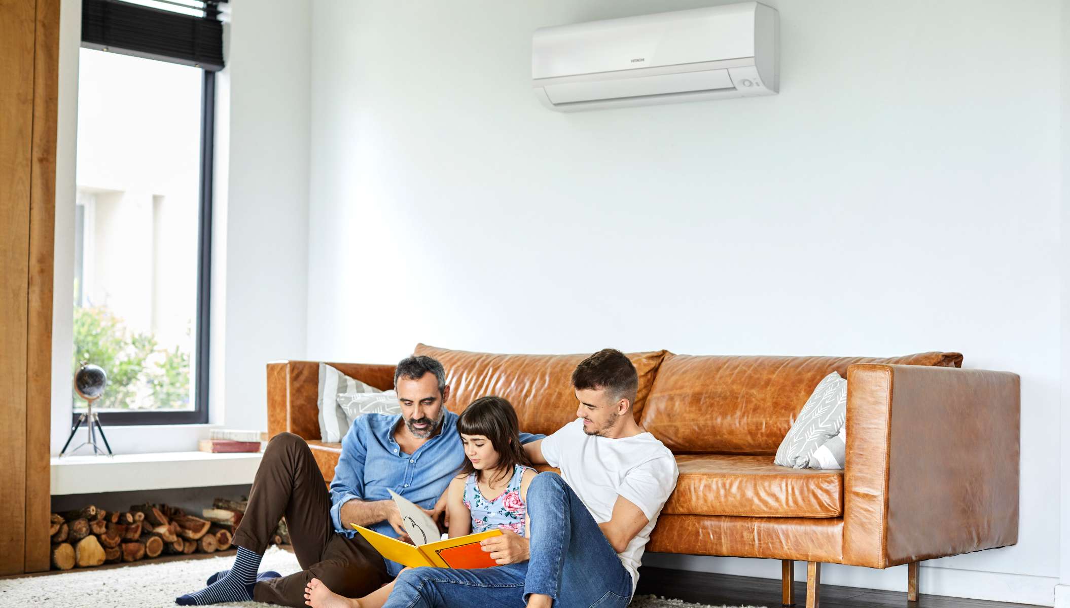 Two fathers and their daughter read a book in the living room, sitting on a rug next to a leather sofa. An indoor air conditioner unit ensures a comfortable temperature.
